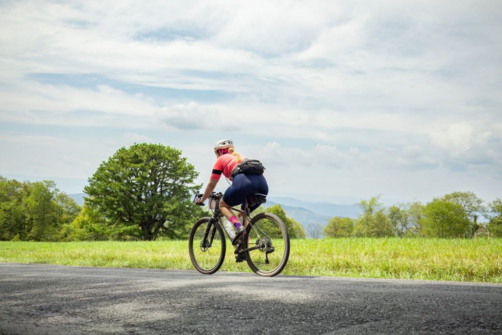 A cyclist rides on Howard's Lick Road near Mathias, WV with a field and mountains in the background.